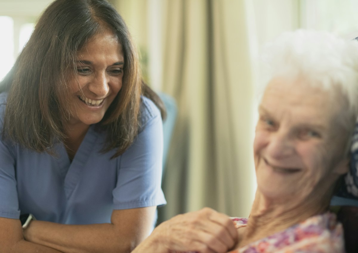 Opiekunka medyczna rozmawia z uśmiechniętą seniorką w jej domu